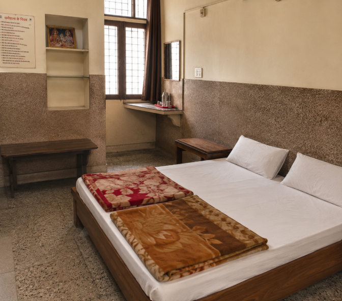 Simple and clean room at Barnala Dharamshala, Haridwar, featuring a wooden bed with fresh linens, traditional blankets, basic furnishings, natural daylight, and essential amenities, offering a comfortable and budget-friendly stay for Kanwar Yatra pilgrims visiting Haridwar. 